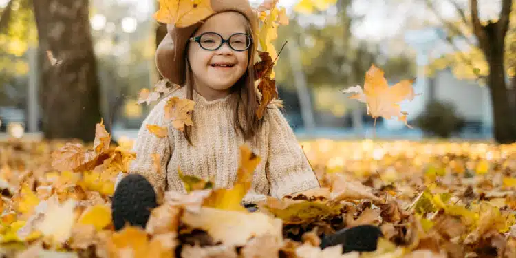 Happy cute brown-haired woman with Downs syndrome in a knitted sweater and stylish glasses throws bright foliage into the sky and laughs, the child enjoys time in a warm autumn park, happy childhood
